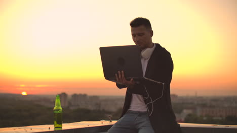A-young-man-sitting-on-the-edge-of-the-roof-with-a-laptop-and-a-beer-working-typing-on-a-laptop